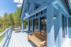 A wooden deck with a bench and a table, surrounded by glass doors and windows, overlooking a forested area.