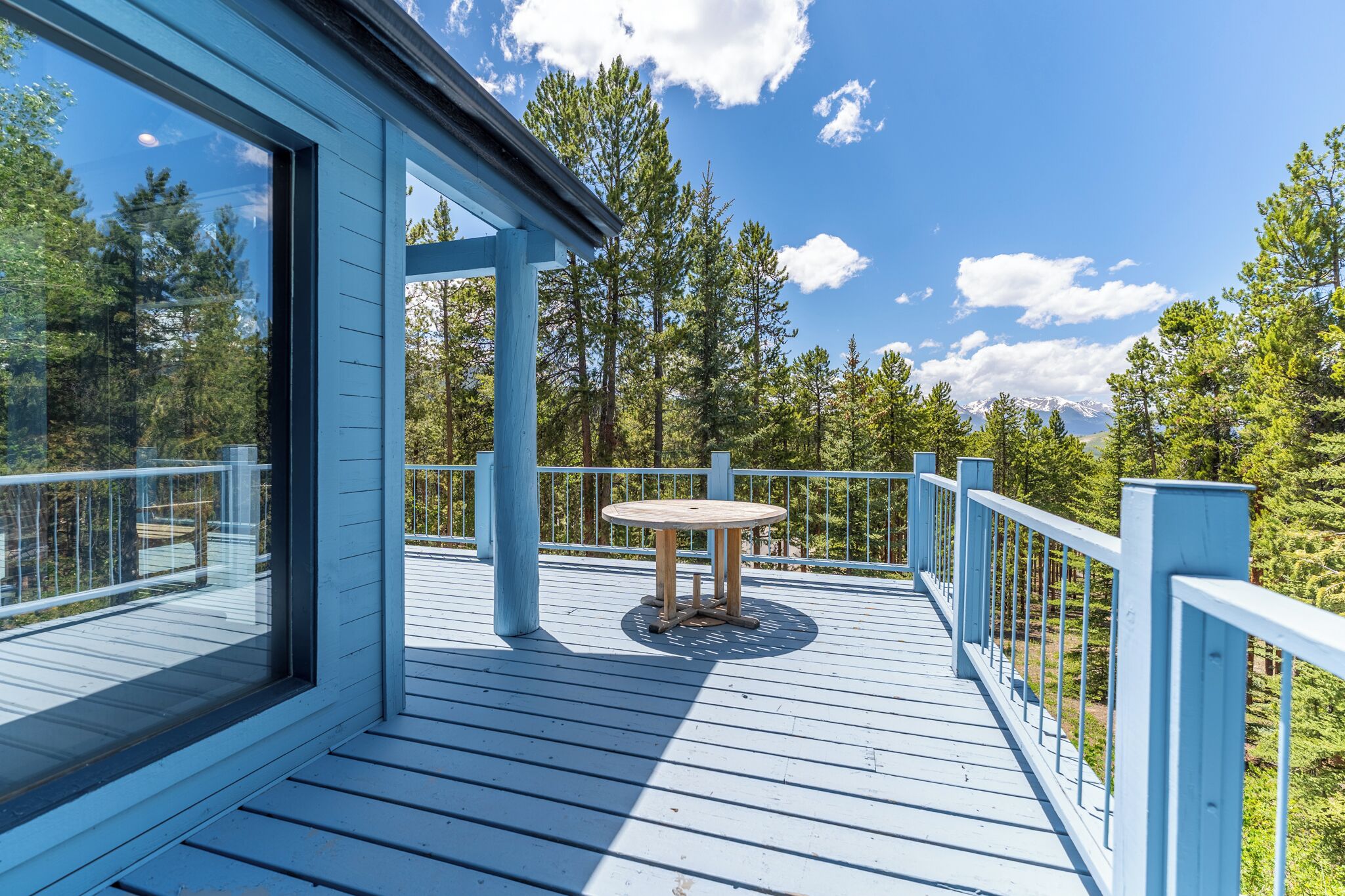 Eine Holzterrasse mit einem runden Tisch und blauen Geländern, umgeben von Bäumen und blauem Himmel.