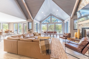 A spacious living room with a large sectional sofa, a fireplace, and a dining area with a high ceiling and large windows.