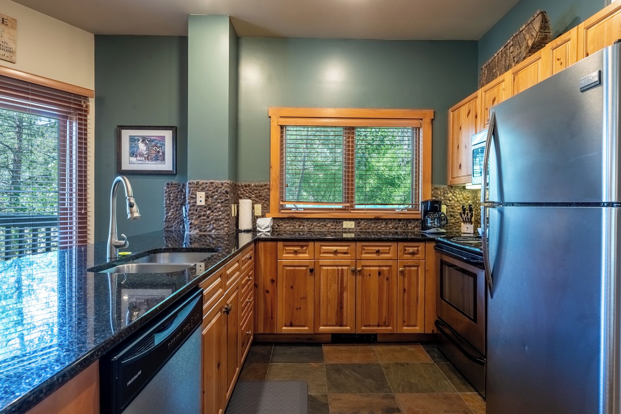 A modern kitchen with wooden cabinets, a stainless steel refrigerator, and a window with a view of trees.