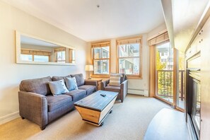 Una sala de estar moderna con sofá, mesa de centro y sillas. Hay ventanas grandes con persianas, un espejo en la pared y vistas a edificios.