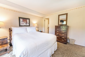 Cama bem arrumada com lençóis brancos, cômoda de madeira com gavetas, quadro emoldurado na parede e duas luminárias de cabeceira.