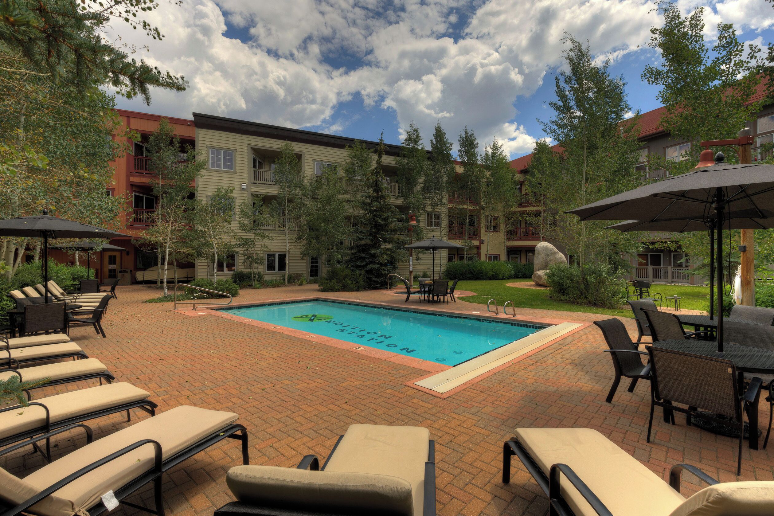 A pool area with lounge chairs and umbrellas, surrounded by buildings and trees.