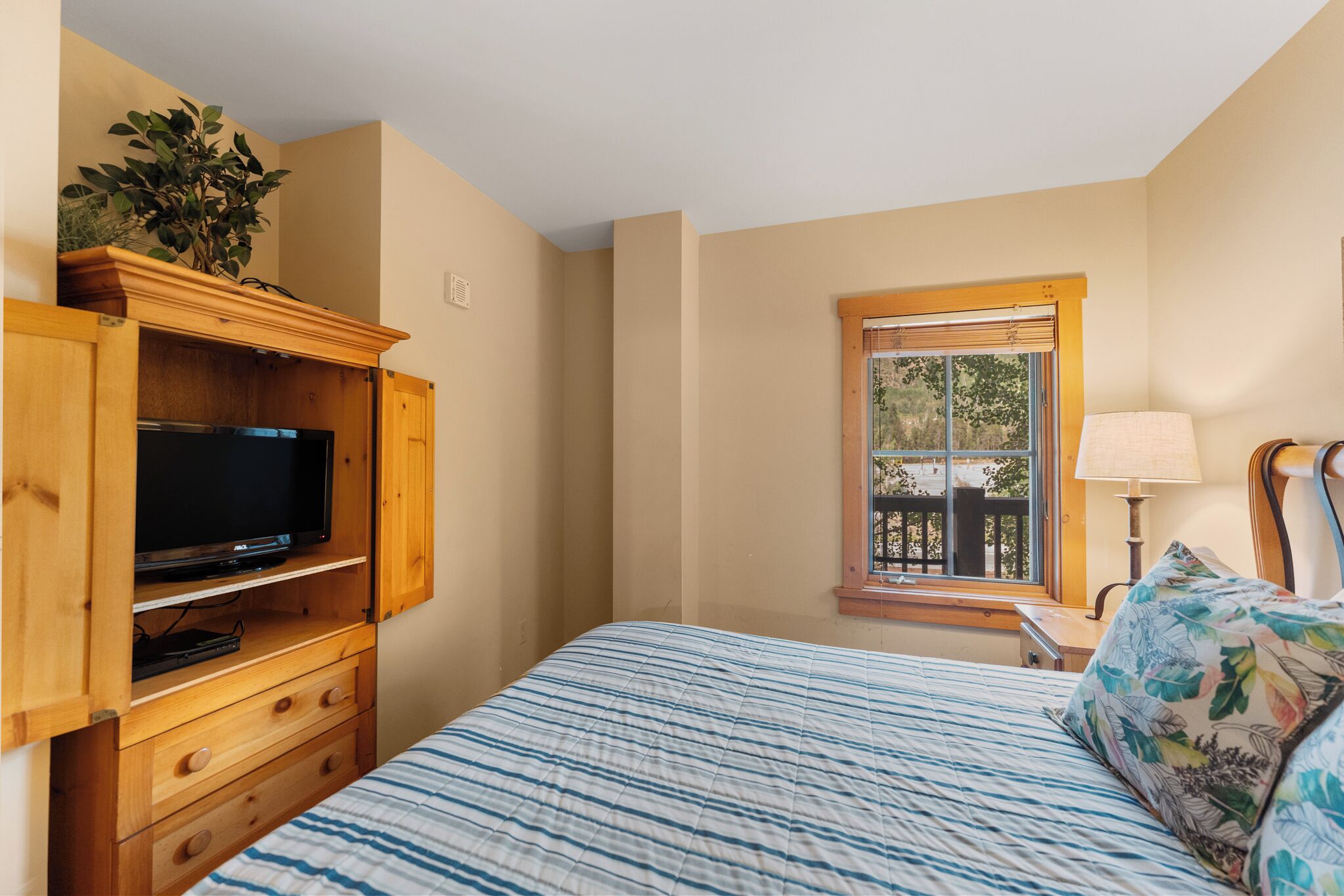 Een slaapkamer met een bed, een televisie op een houten standaard, een raam met uitzicht en een nachtkastje met een lamp.