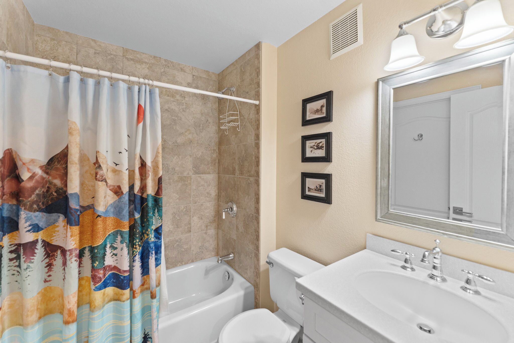 Une salle de bain équipée d’une baignoire, de toilettes, d’un lavabo et d’un rideau de douche orné d’un motif naturel. Trois tableaux encadrés sont accrochés au mur.