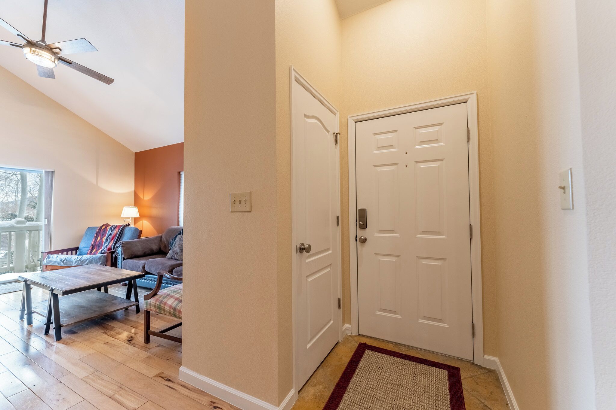 A hallway with a white door, a ceiling fan, a wooden table, and a sofa with a colorful blanket.