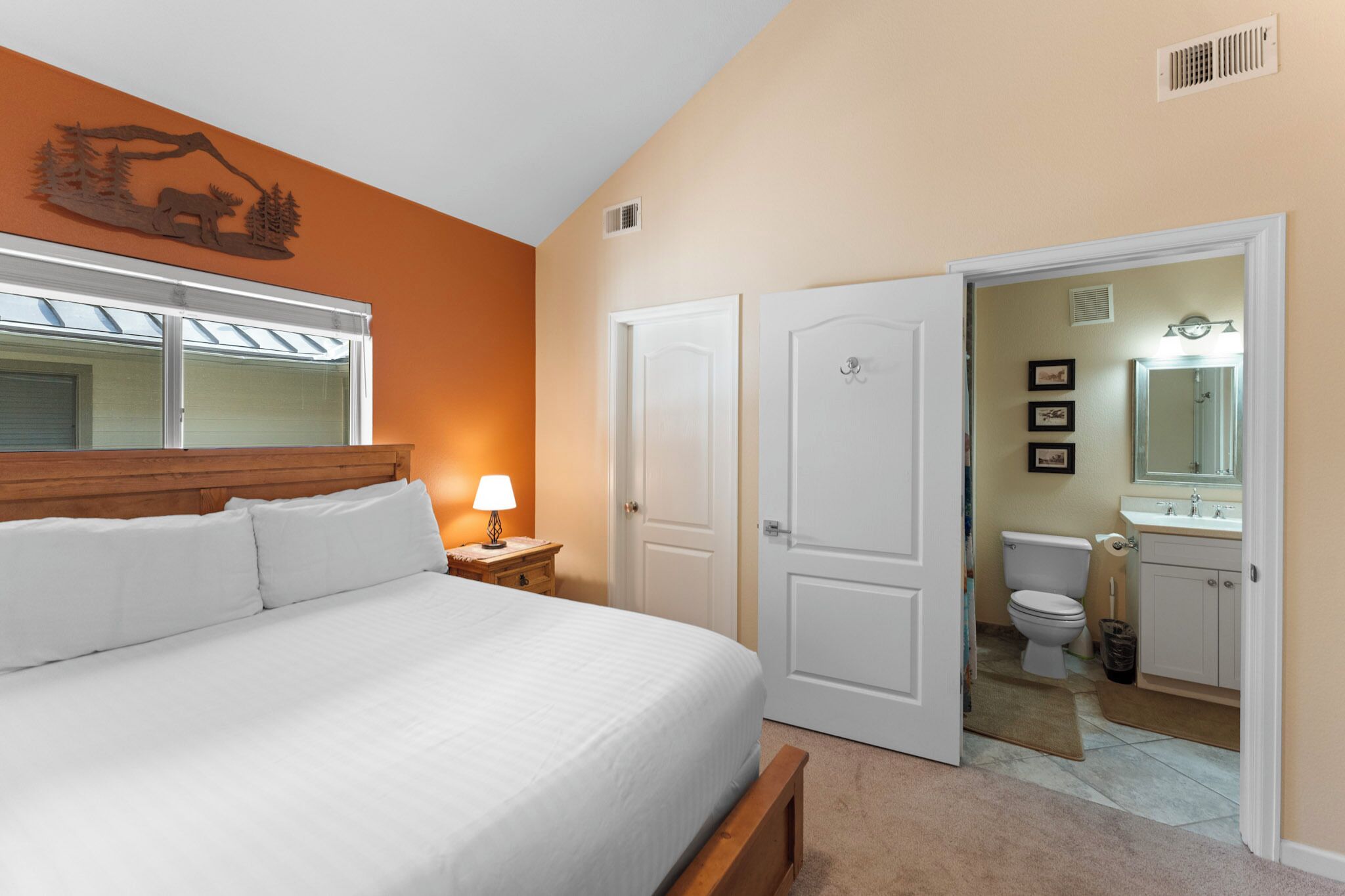 A bedroom with a bed, nightstand, and lamp. There is a door leading to a bathroom with a toilet and sink.