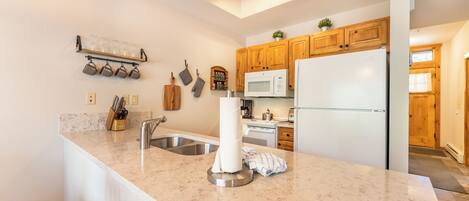 A kitchen with a marble countertop, stainless steel sink, wooden cabinets, a microwave, and a refrigerator.