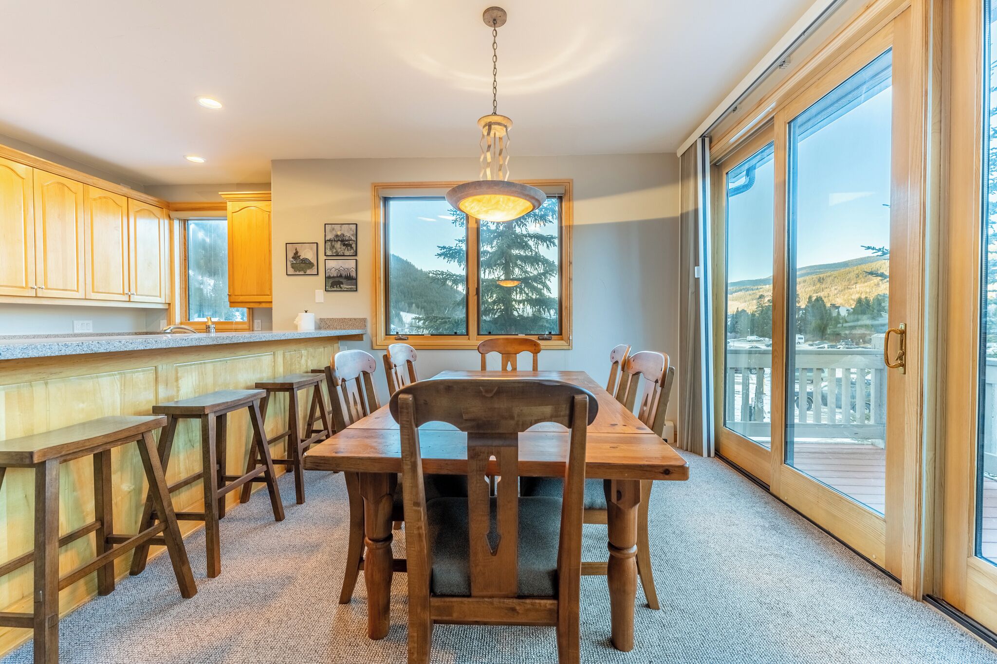 Un comedor con mesa y sillas de madera, ventanas amplias con vistas al exterior y una lámpara colgante.
