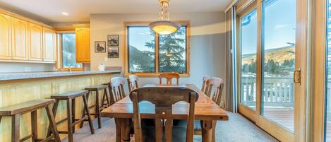 Un comedor con mesa y sillas de madera, ventanas amplias con vistas al exterior y una lámpara colgante.