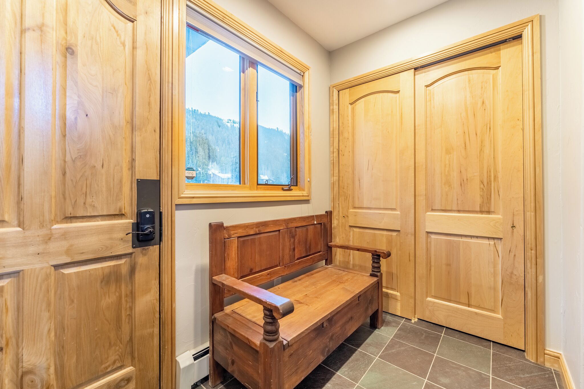 A wooden bench in a corner with two wooden doors and a window with a view of a forest.