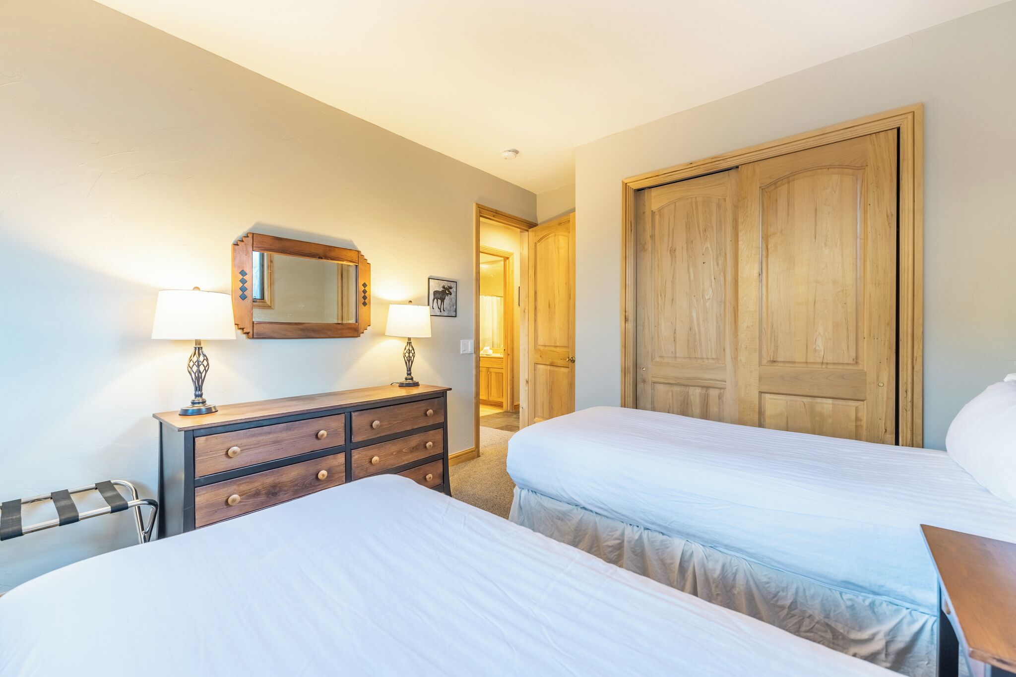 A hotel room with two beds, a wooden wardrobe, a dresser with drawers, a mirror, and a lamp.