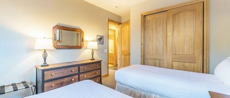 A hotel room with two beds, a wooden wardrobe, a dresser with drawers, a mirror, and a lamp.