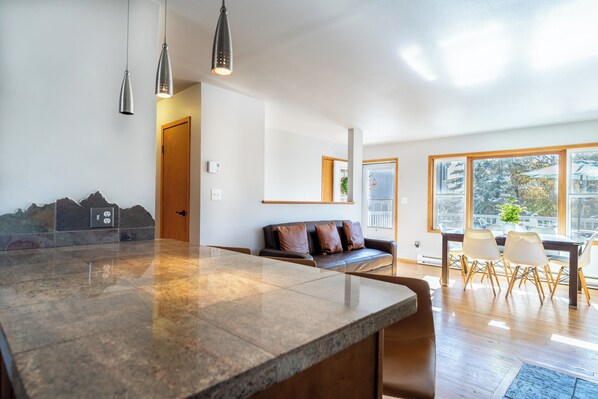 Una cocina moderna con una isla grande, un comedor con mesa y sillas de madera, y un rincón de estar con sofá y cojines.