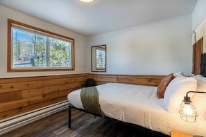 Un dormitorio con suelo de madera, una cama con ropa de cama blanca, una manta verde, una silla negra, una mesita de noche con una lámpara y un ventanal con vistas a árboles y una valla.