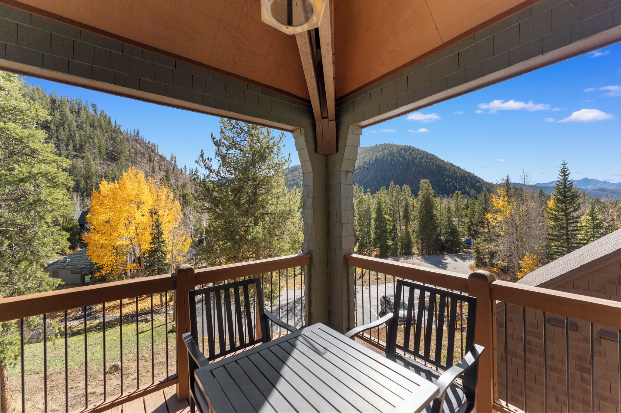 A wooden deck with a table and chairs, overlooking a forested area with autumn foliage and distant mountains.