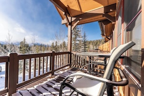 A wooden deck with a table and chairs, overlooking a snowy landscape with trees and mountains.