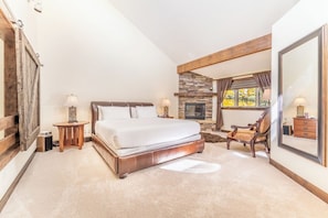 A bedroom with a large bed, a fireplace, a wooden ceiling, and a window with curtains.