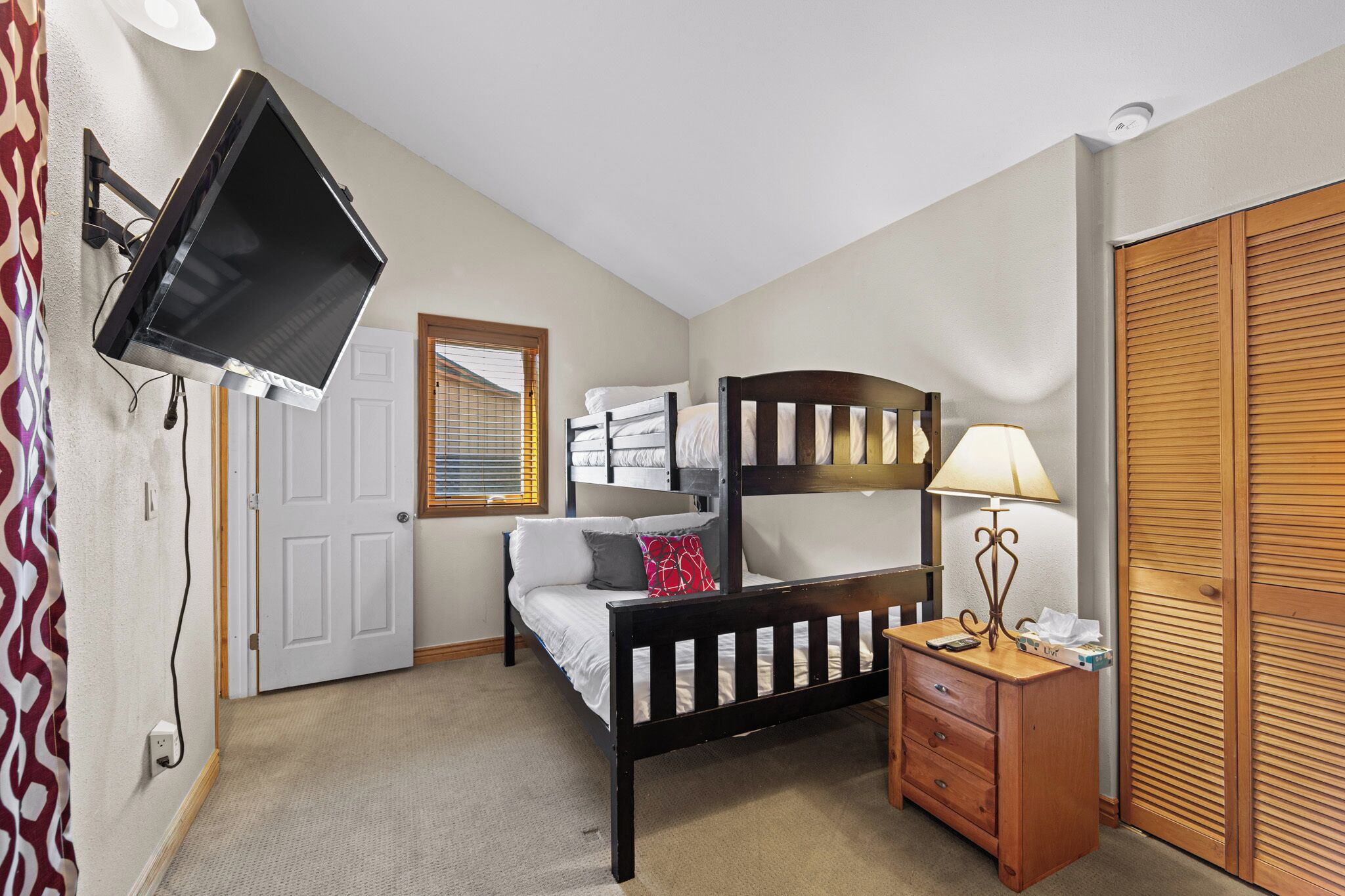 A bedroom with a bunk bed, a bedside table with a lamp, a TV mounted on the wall, and a wardrobe.