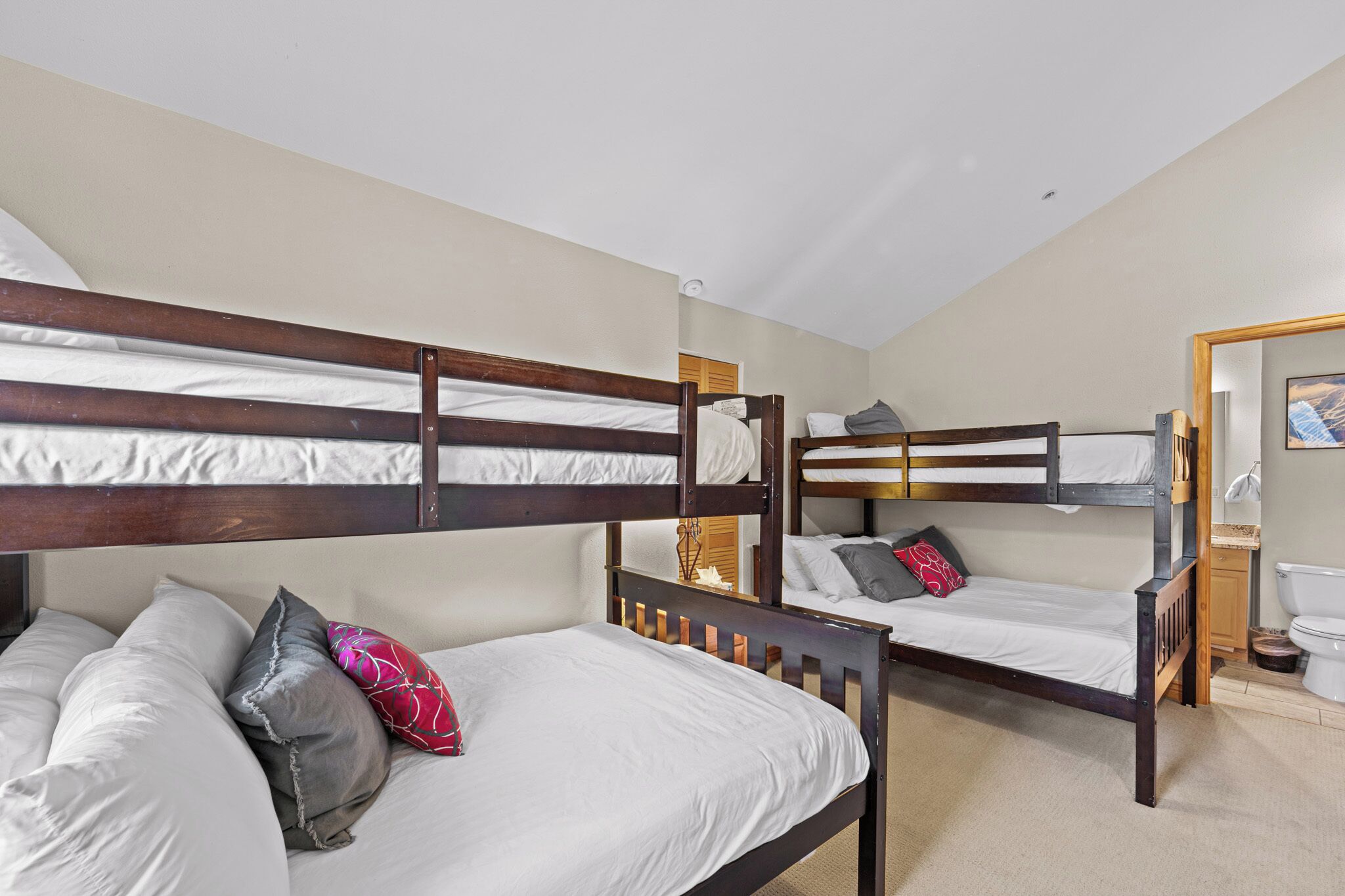 A room with three bunk beds, each with white bedding and pillows, and a wooden headboard. A small bathroom with a toilet and sink is visible to the right.