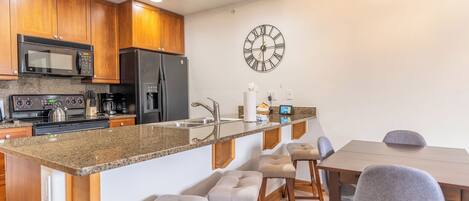 Una cocina moderna con armarios de madera, un refrigerador de acero inoxidable, un microondas y un horno empotrado. Se observa un reloj de pared grande y una zona con taburetes, una mesa y sillas de madera.