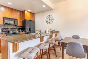 A modern kitchen with wooden cabinets, a stainless steel refrigerator, a microwave, and a built-in oven. There is a granite countertop, a sink, and bar stools.