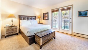 Un dormitorio con una cama grande, dos mesitas de noche con lámparas, un banco de madera y una puerta corrediza de vidrio que da a un área exterior.