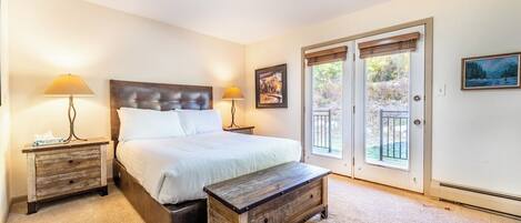 A bedroom with a large bed, two bedside tables with lamps, a wooden bench, and a sliding glass door leading to an outdoor area.