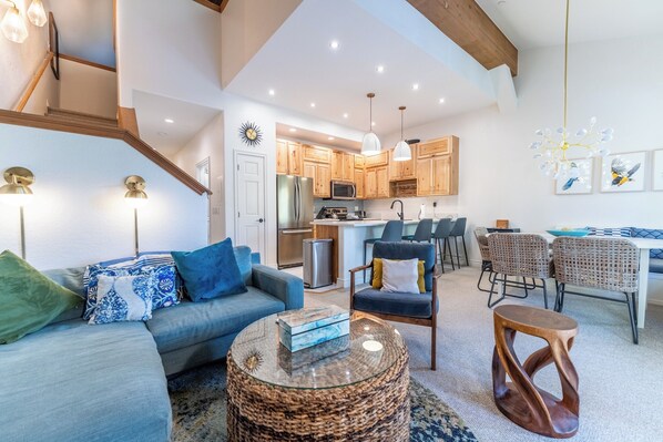 A modern living room with a sofa, armchair, and coffee table, a dining area with wicker chairs, and a kitchen with wooden cabinets and a central island.