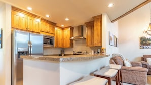 A kitchen with wooden cabinets, a stainless steel refrigerator, a built-in oven, and a bar stool area. - Ab22-3 (Keystone)