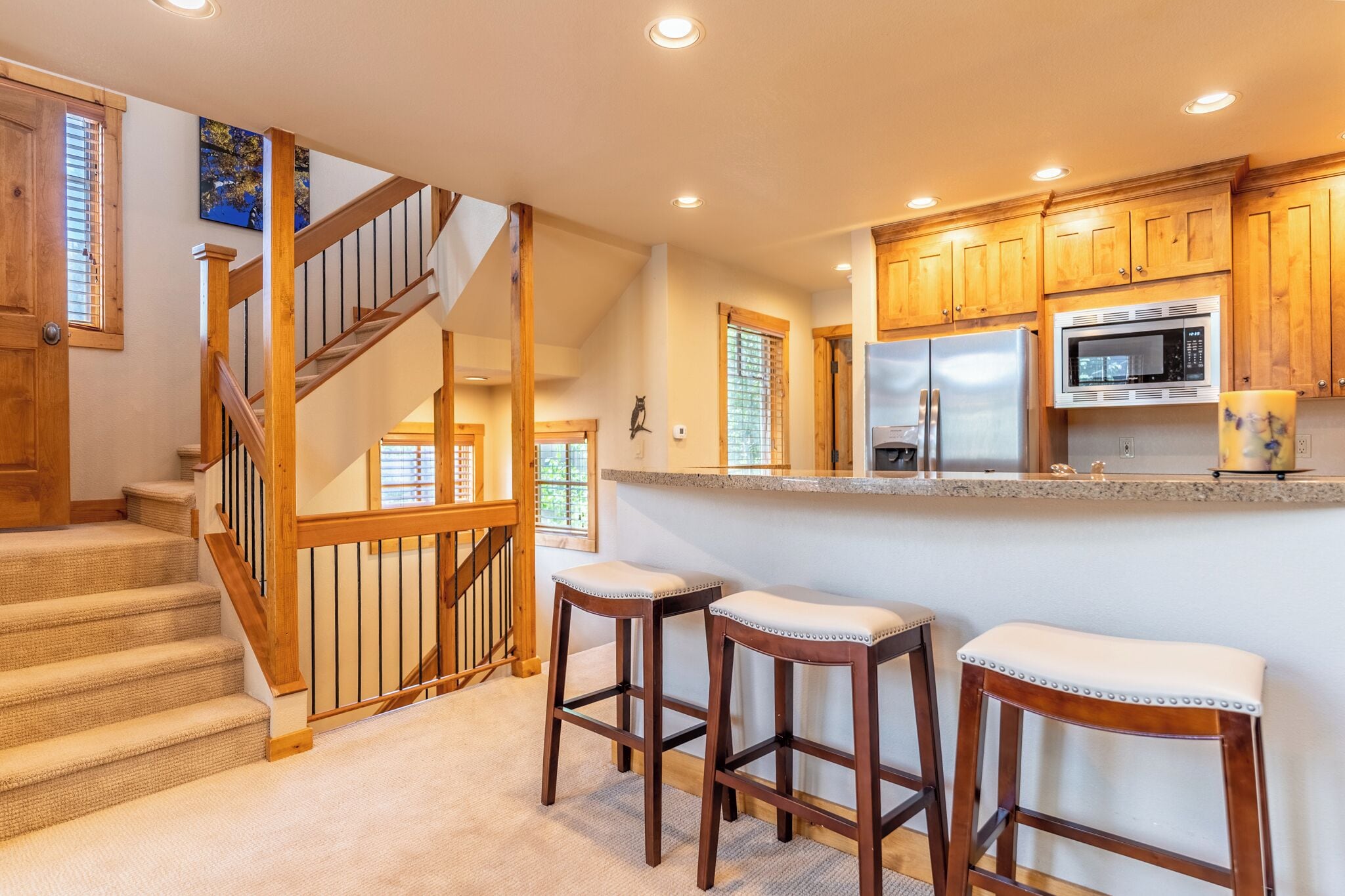 A kitchen with wooden cabinets, a stainless steel refrigerator, a microwave, and a granite countertop. There are two wooden stools with cushioned seats.