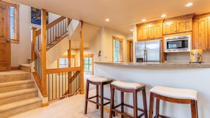 A kitchen with wooden cabinets, a stainless steel refrigerator, a microwave, and a granite countertop. There are two wooden stools with cushioned seats. - Ab22-3 (Keystone)