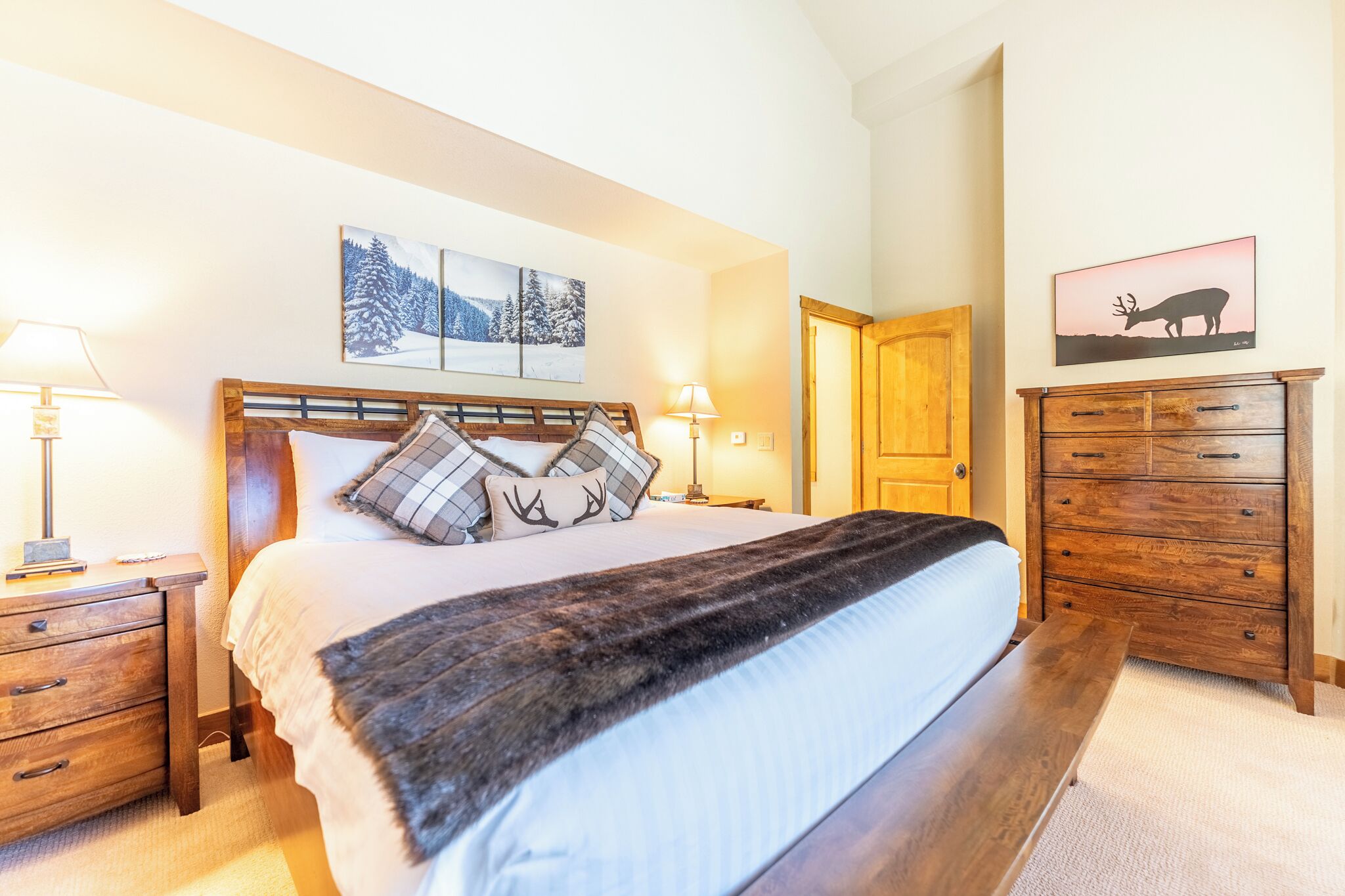 A bedroom with a large bed, wooden headboard, two bedside tables with lamps, a wooden dresser, and two wall-mounted paintings.