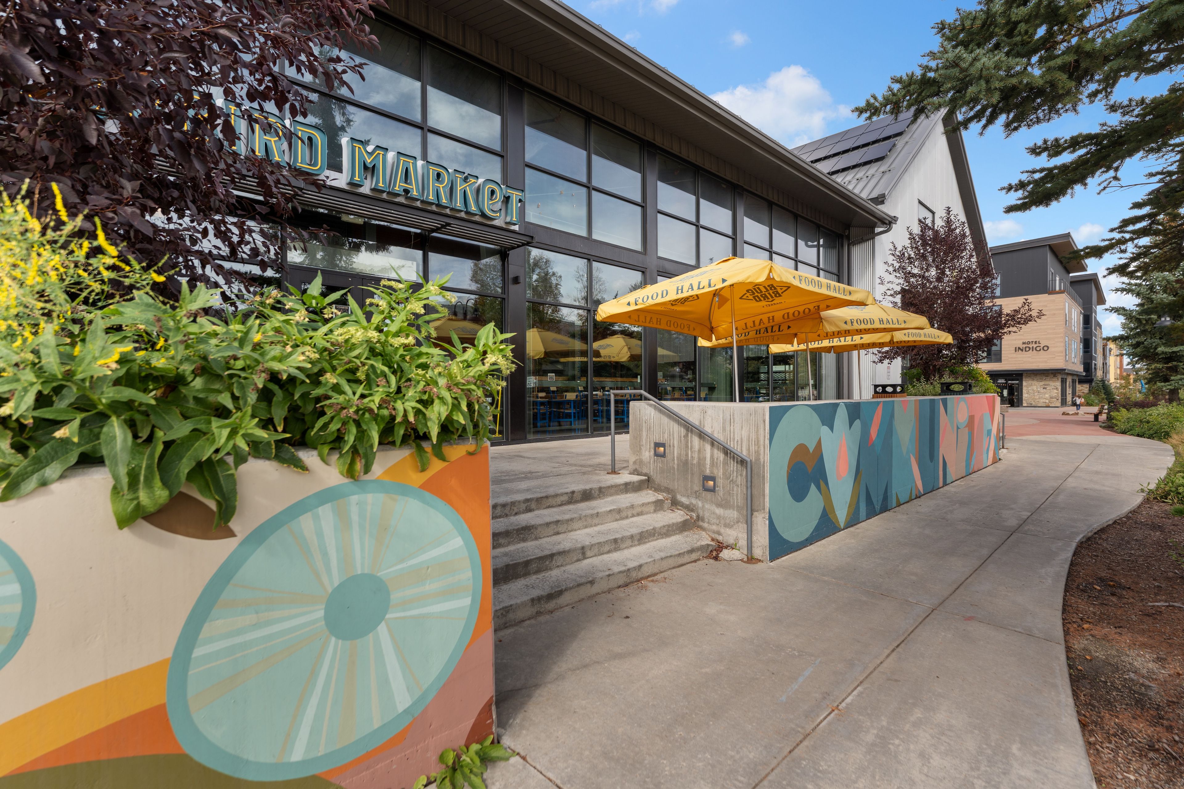 A building with a sign that reads 'MARKET' and 'INDIGO HOTEL' on the side. There are outdoor umbrellas and a colorful mural on the wall.