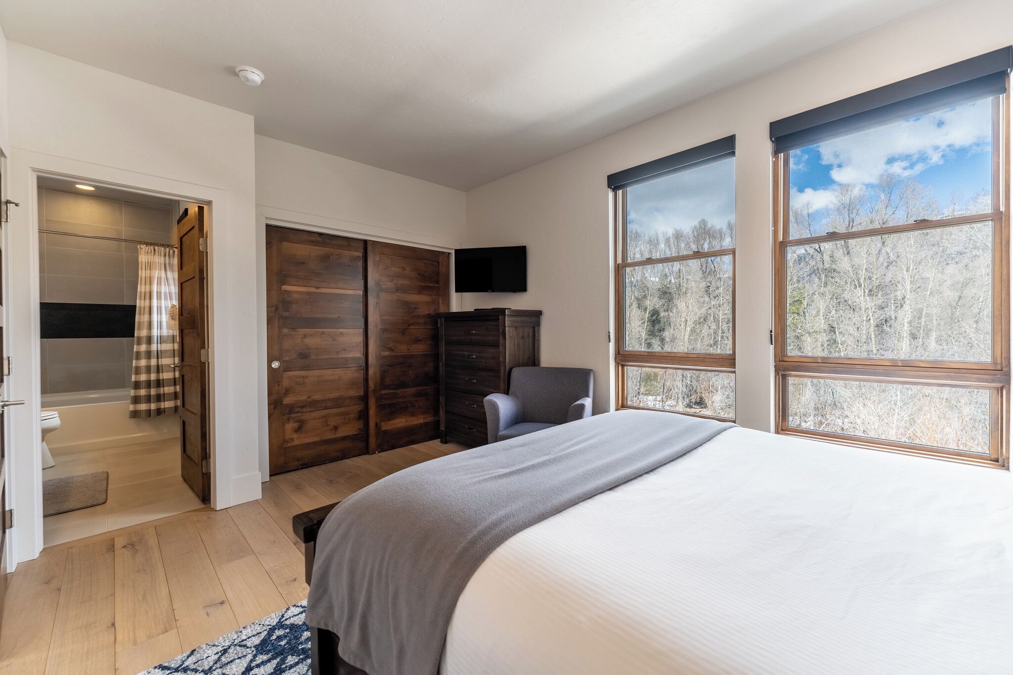 A bedroom with a large bed, a wooden wardrobe, a television, and a window with a forest view.