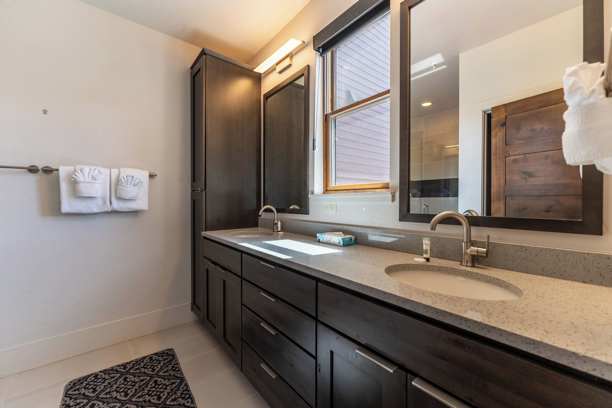 A modern bathroom with two sinks, dark cabinetry, a large mirror, and a window.