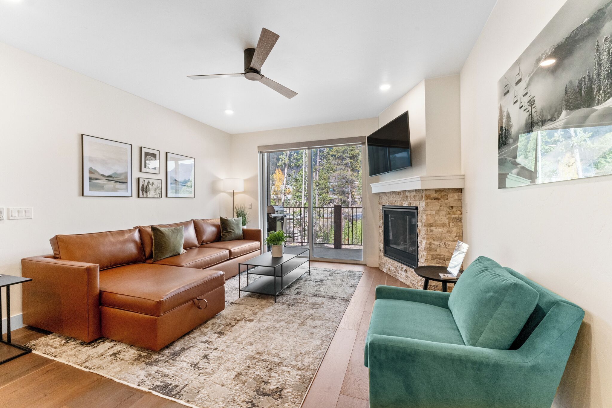 A modern living room with a brown leather sofa, a green armchair, a fireplace, and a large window with a view of trees.