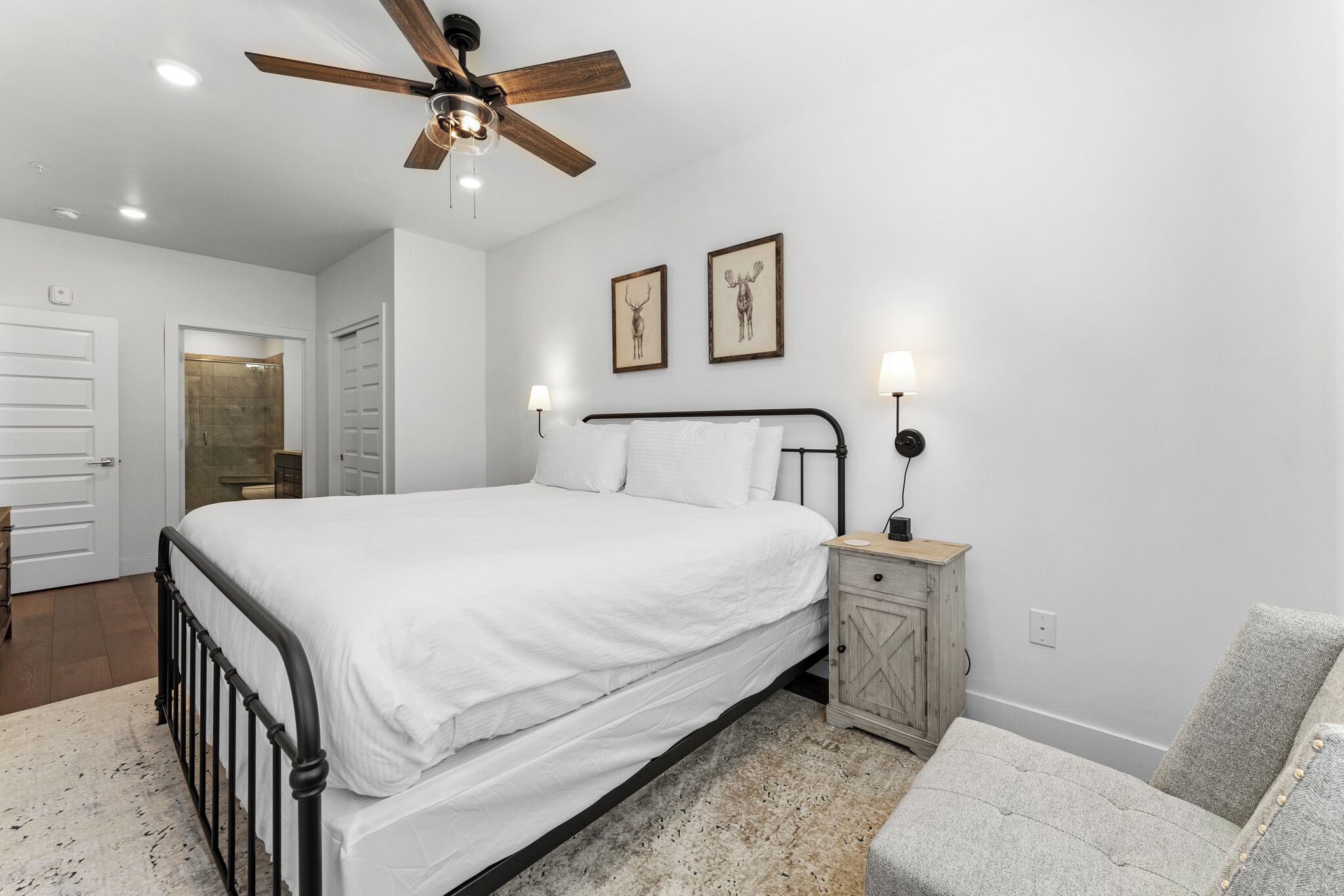 A bedroom with a bed, a nightstand, a chair, a ceiling fan, and two framed pictures on the wall.