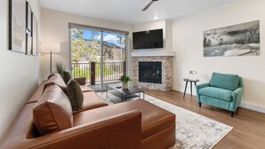 A modern living room with a leather sofa, a teal armchair, a fireplace, and a large window offering a view of the outdoors. - Clearwater Lofts #203, Building 2 (Keystone)