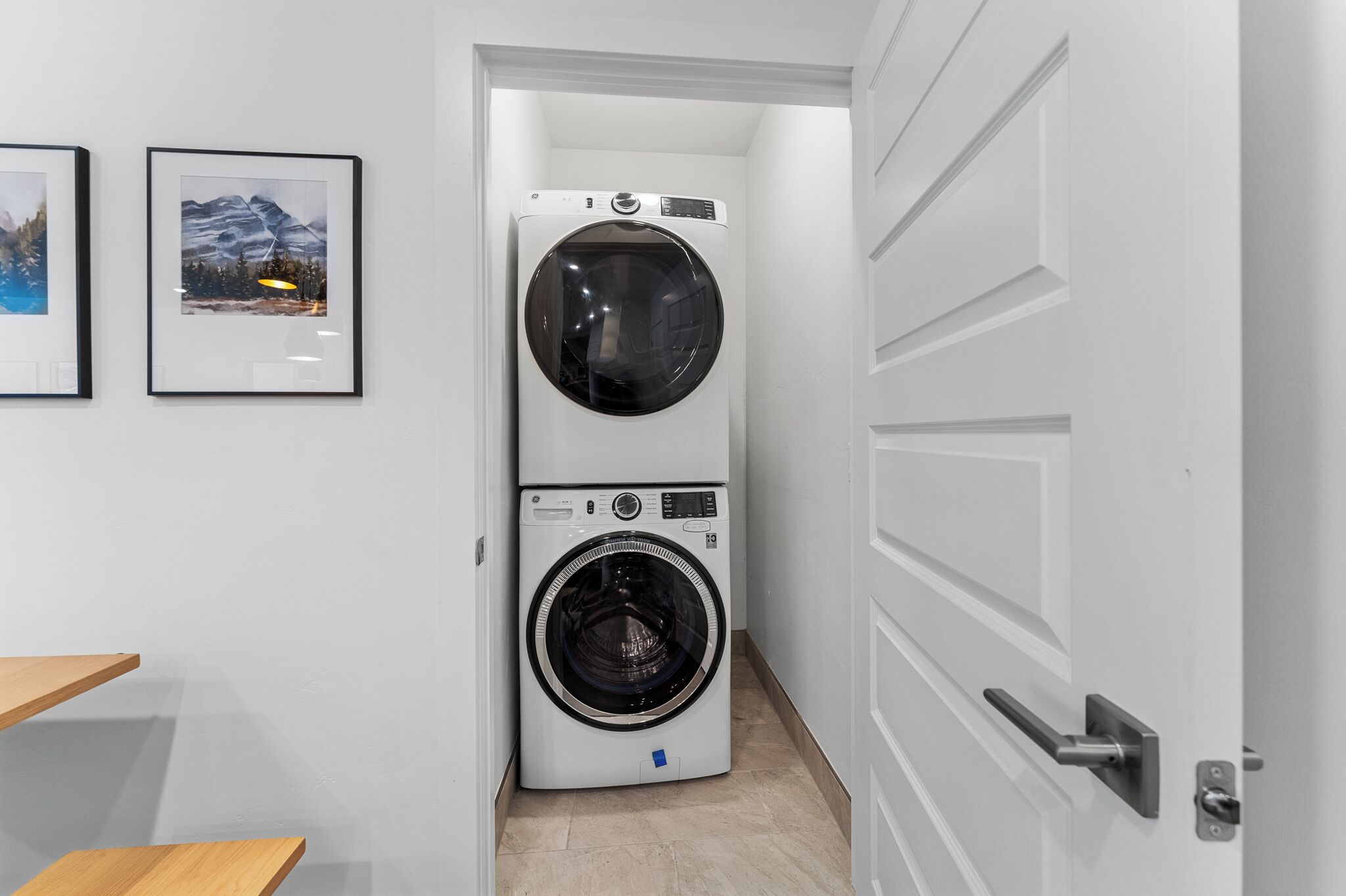 A laundry room with two stacked washing machines, a wooden bench, and framed pictures on the wall.