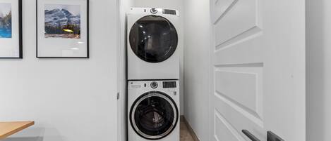 A laundry room with two stacked washing machines, a wooden bench, and framed pictures on the wall.
