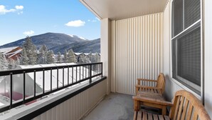 Un balcón con una mesa y sillas de madera, vista de montañas nevadas y un edificio al fondo.