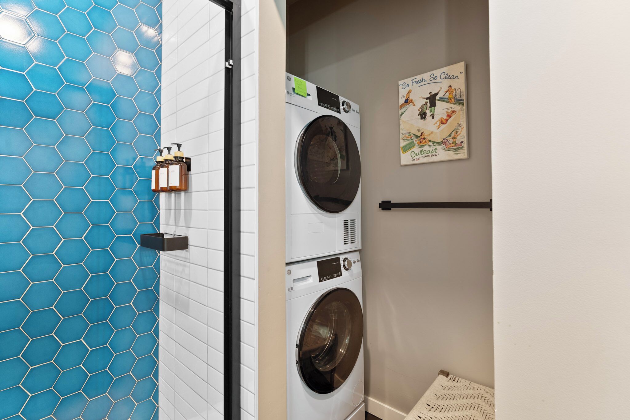 A laundry area with a stacked washer and dryer, a blue hexagonal tile wall, and a poster on the wall.