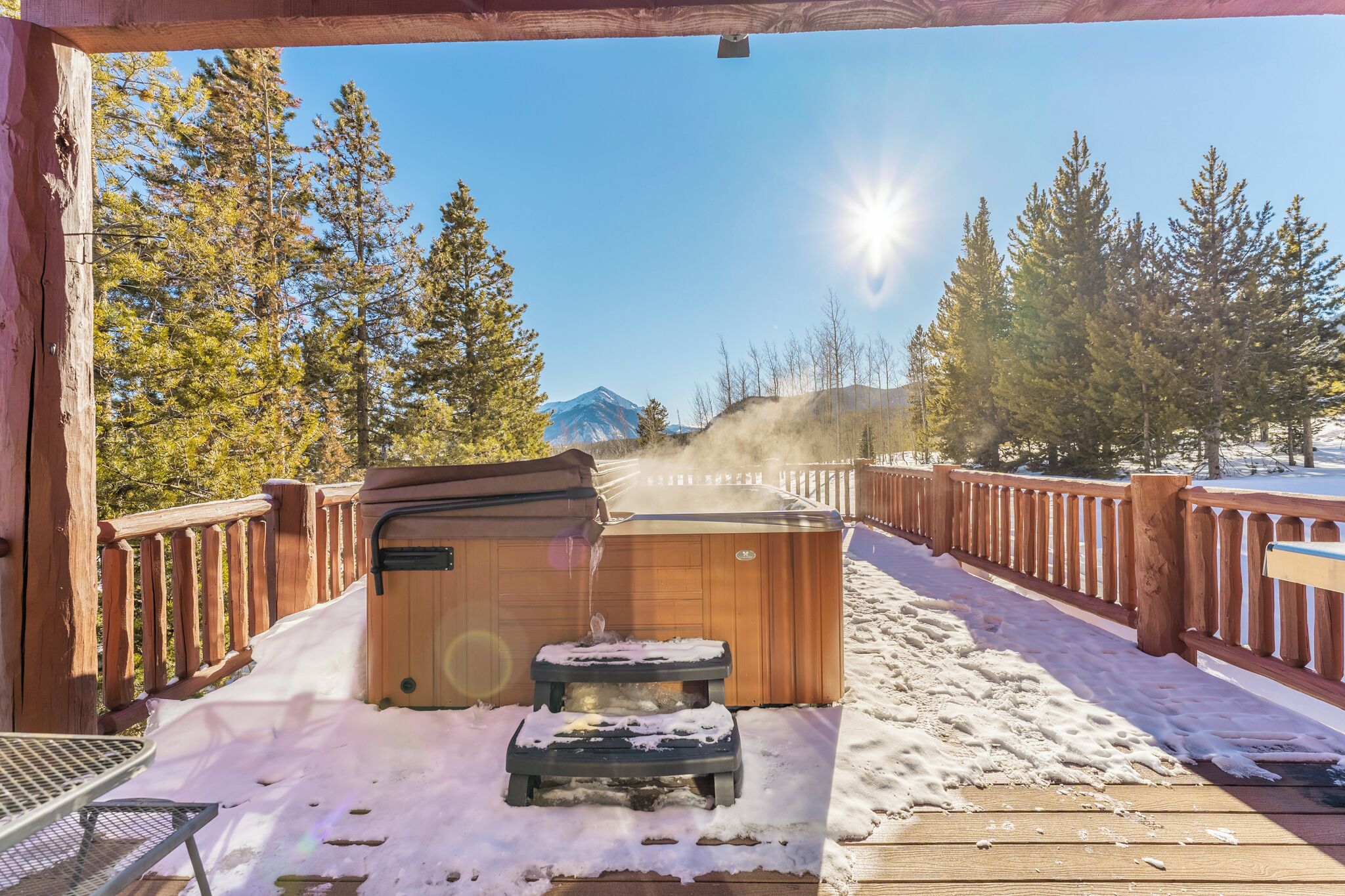 A wooden deck with a hot tub, surrounded by snow and trees, with a mountain in the background.