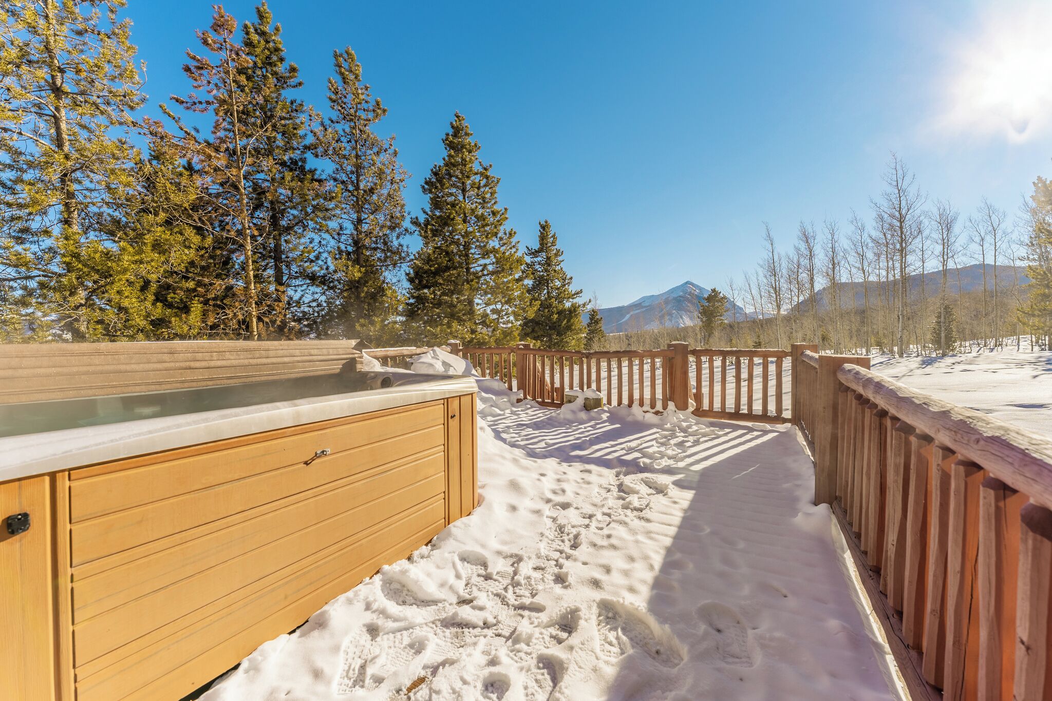 A wooden deck with a glass enclosure, surrounded by snow-covered ground and trees, leading to a mountain in the background.
