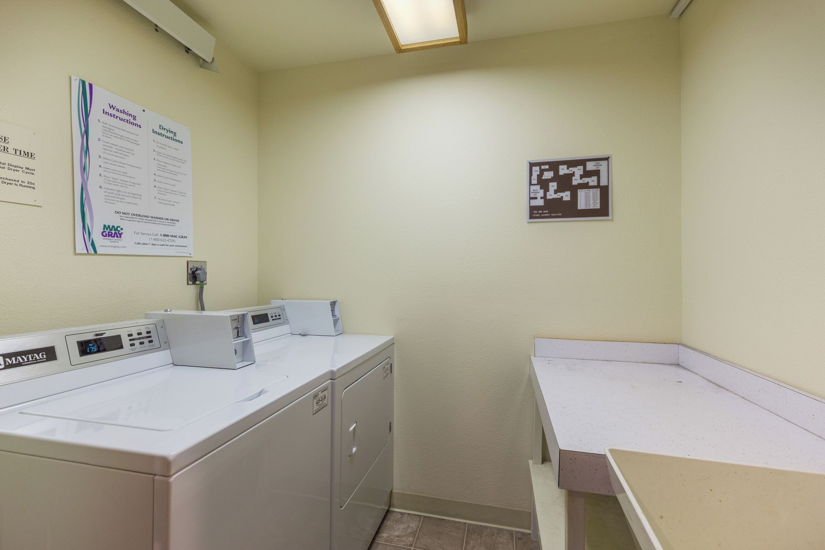 Un cuarto de lavandería con lavadora y secadora Maytag de uso comercial, un letrero de instrucciones en la pared y un cartel con el plano de distribución.