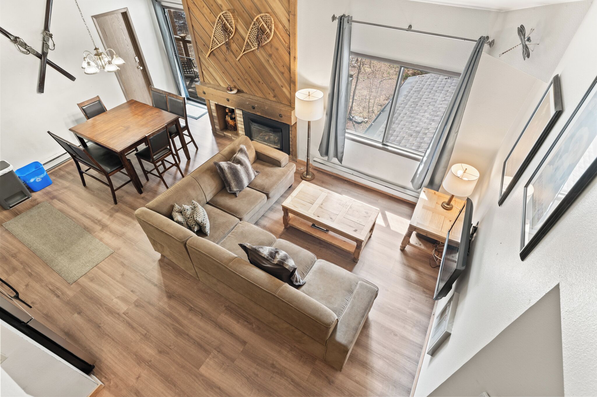 A modern living room with a wooden dining table, sofa, and coffee table, a fireplace, and large windows.