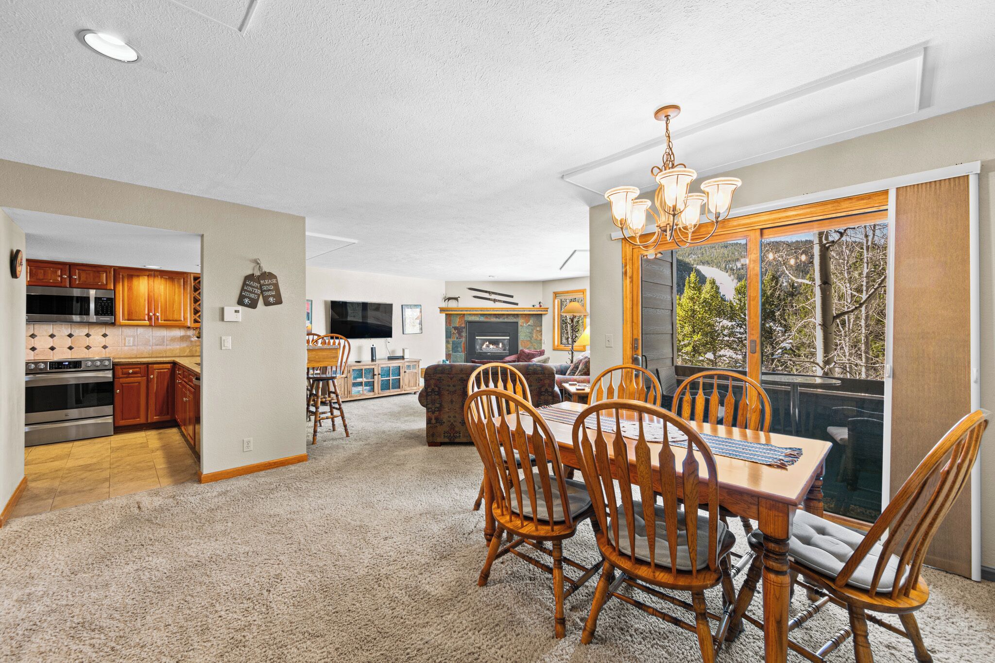 A spacious living area with a dining table, chairs, and a chandelier. There is a kitchen area with wooden cabinets and a large window overlooking a snowy landscape.
