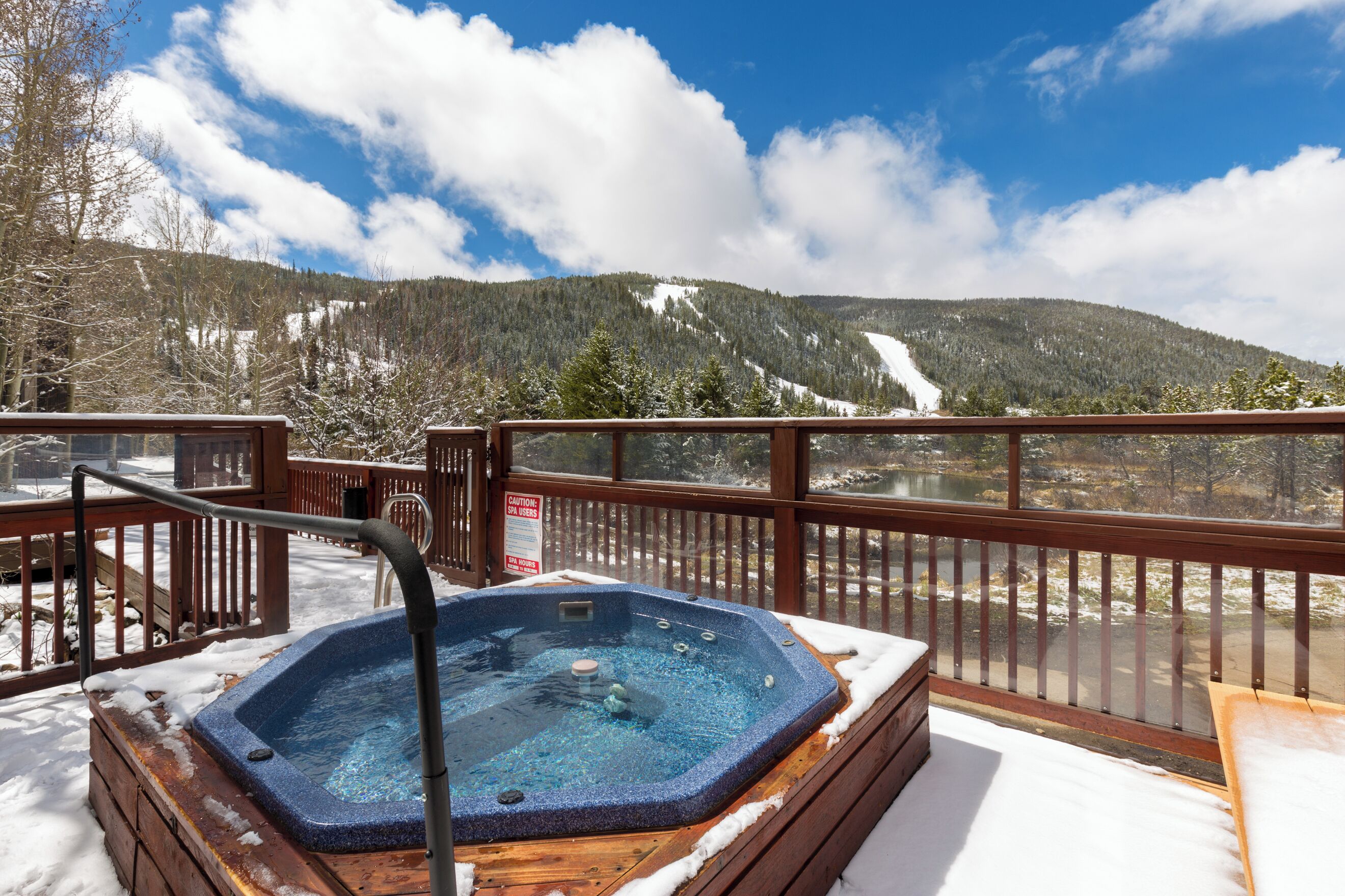 A hot tub on a wooden deck with a view of a snowy mountain and a lake.