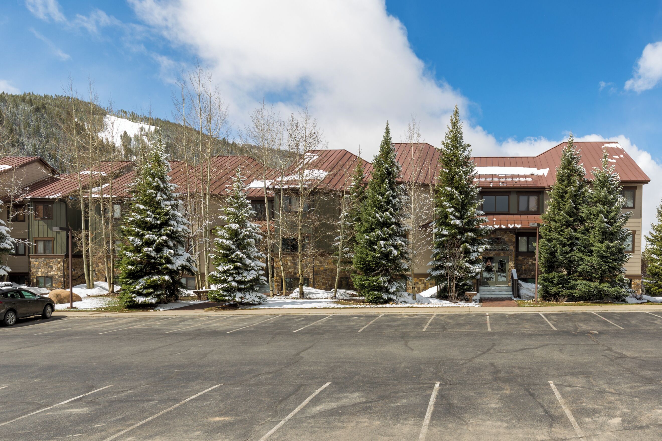 A hotel with a parking lot, snow-covered trees, and a mountain backdrop.